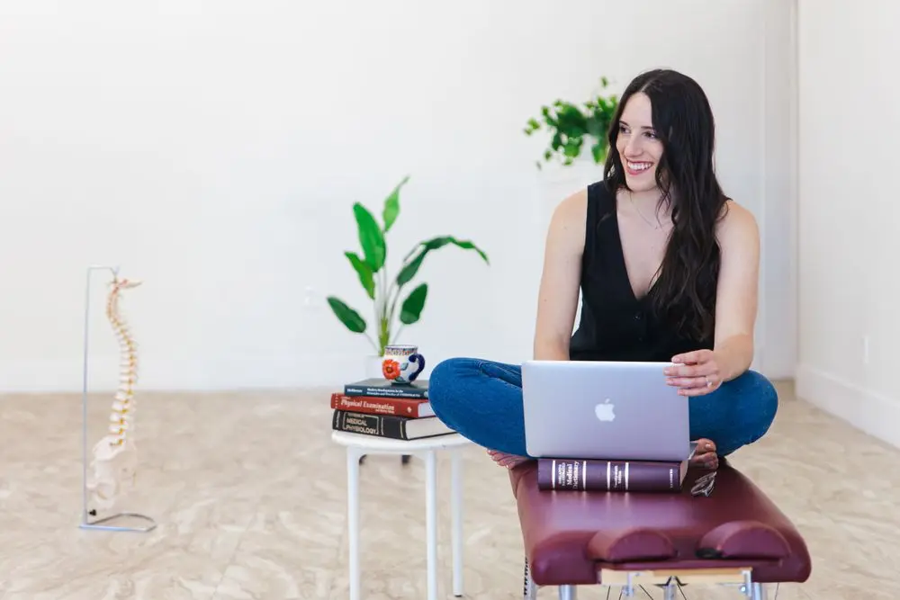 Woman smiling off-camera with a laptop, seated cross-legged on a treatment table at the right of a wide frame, open white wall on the left, photographed by Create With Amy