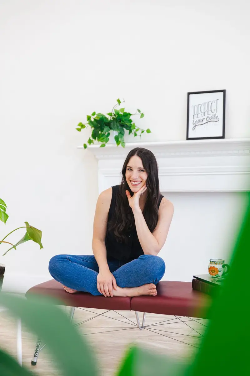 Woman laughing with her hand at her cheek, seated cross-legged and barefoot on a treatment table in her own bright office, a plant soft in the foreground, photographed by Create With Amy