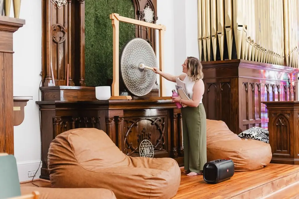 Woman in green wide-leg trousers striking a large gong in an ornate wood-paneled room with organ pipes, notebooks under one arm, photographed by Create With Amy