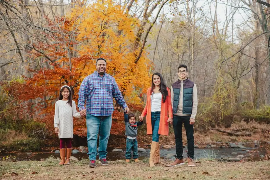 Family of five by a creek under orange fall trees, wearing blue plaid, an orange cardigan, a navy vest, cream and denim, photographed by Create With Amy