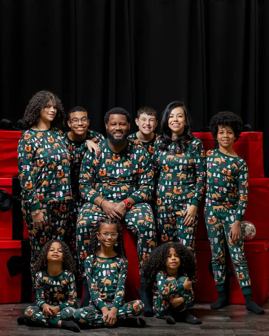 Family of nine in matching dark green holiday pajamas with a woodland animal print, posed on red gift boxes against black drapes, photographed by Create With Amy