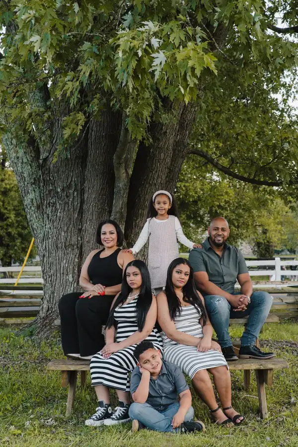 Family of six on and around a wooden bench under a large tree, dressed in black, white, black and white stripes, gray and denim, with one little girl in a pale pink dress, photographed by Create With Amy