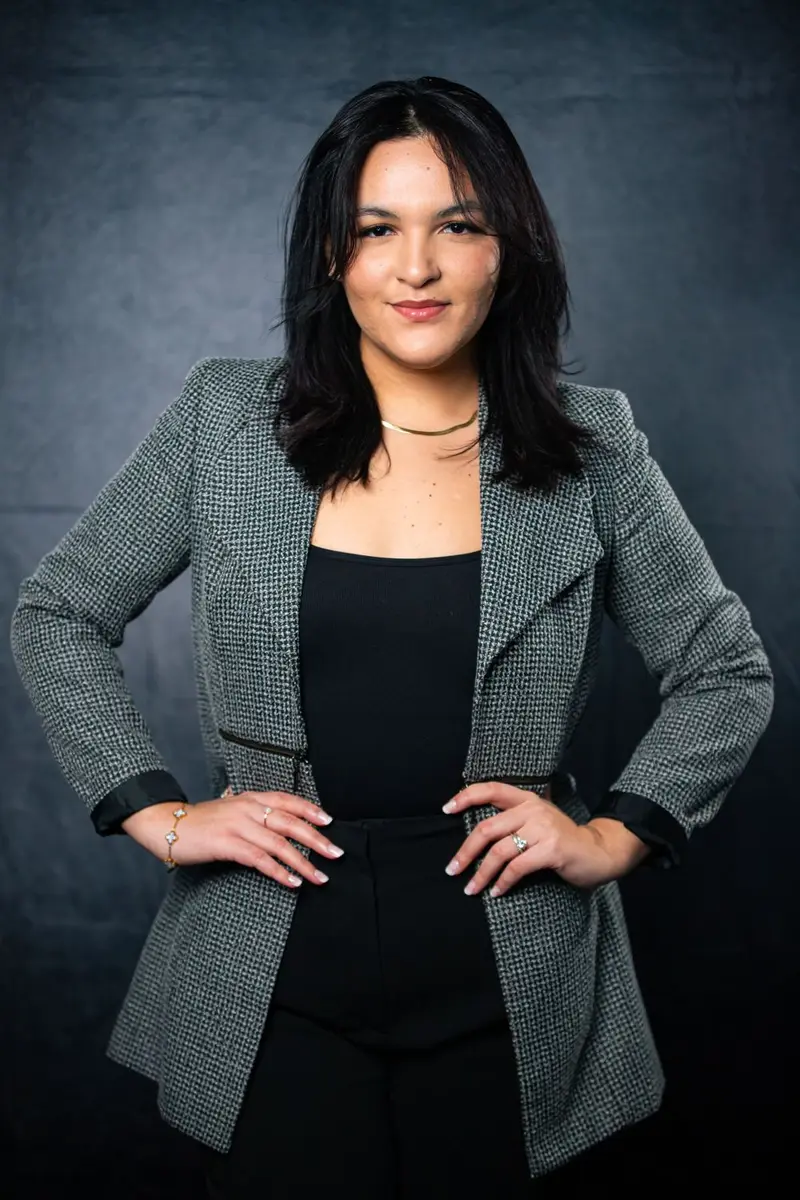 Woman in a gray houndstooth blazer over a black top, hands on her hips, smiling slightly against a dark textured backdrop, photographed by Create With Amy