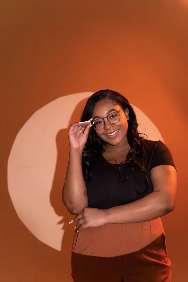 Smiling woman in glasses with her fingertips at the frames, standing in front of a rust backdrop with a circle of light behind her, photographed by Create With Amy