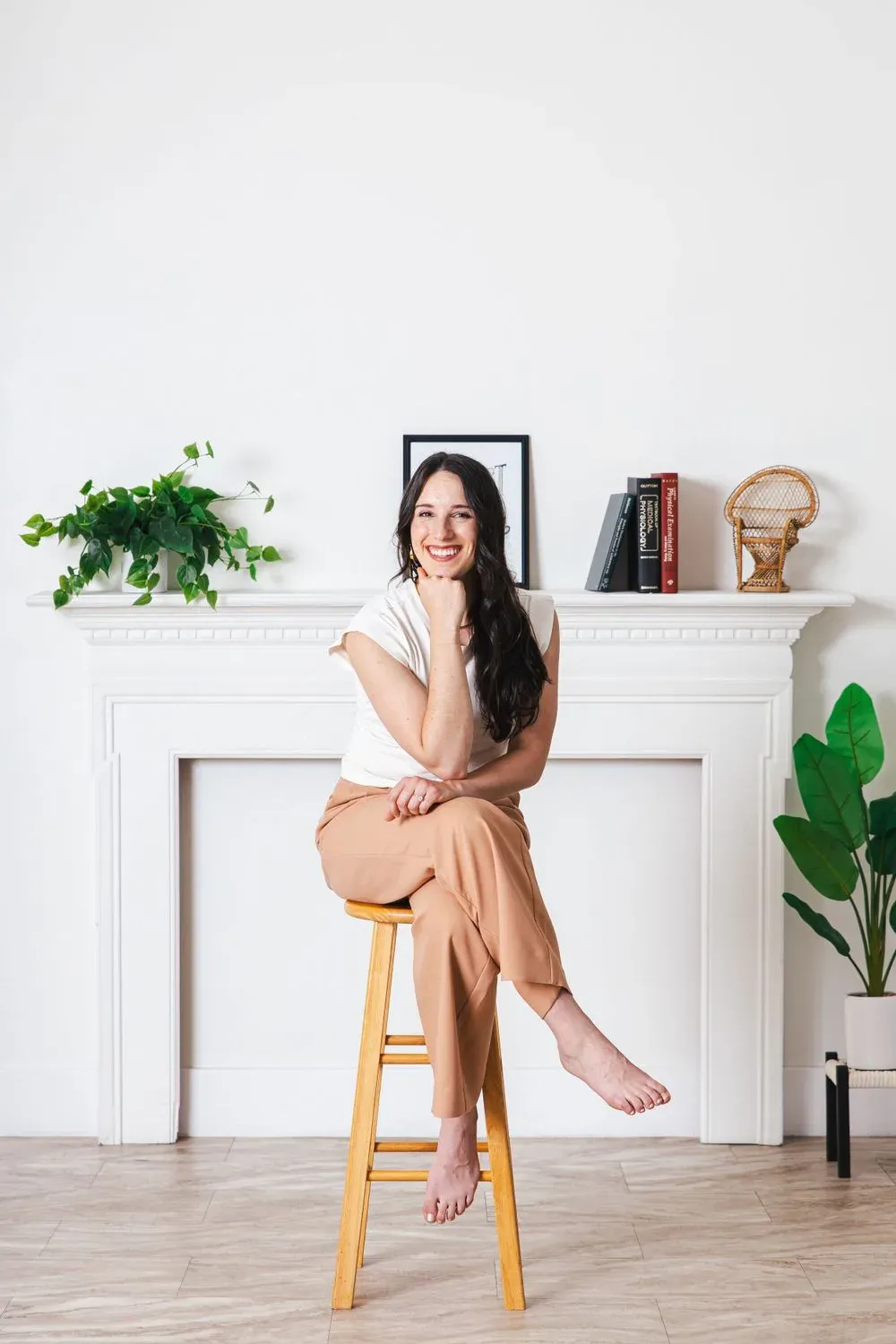 Woman in a cream top and tan wide-leg trousers sitting barefoot on a wooden stool in front of a white fireplace mantel, photographed by Create With Amy