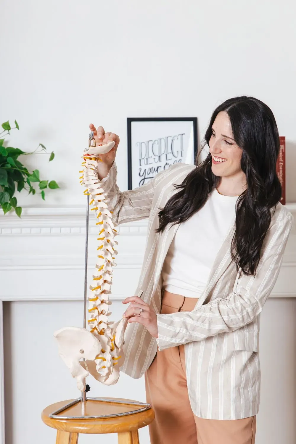 Woman smiling in a cream and tan striped blazer over a white top, holding an anatomical spine model, photographed by Create With Amy