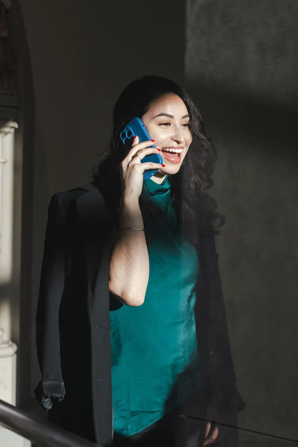 Woman laughing on a phone call in a solid teal top with a black blazer over her shoulders, photographed by Create With Amy