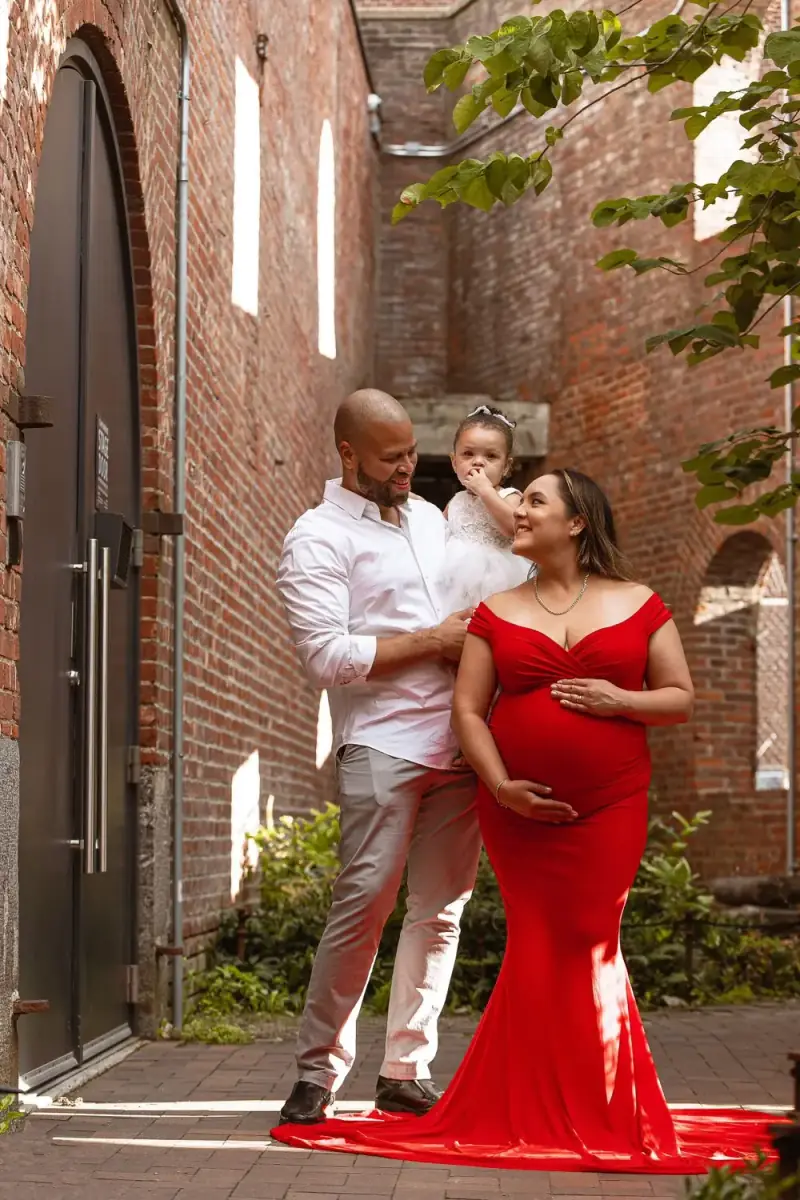 Expecting mother in a red gown smiling up at her partner, who holds their little one in a white lace dress, in a brick courtyard, a family maternity session by Create With Amy