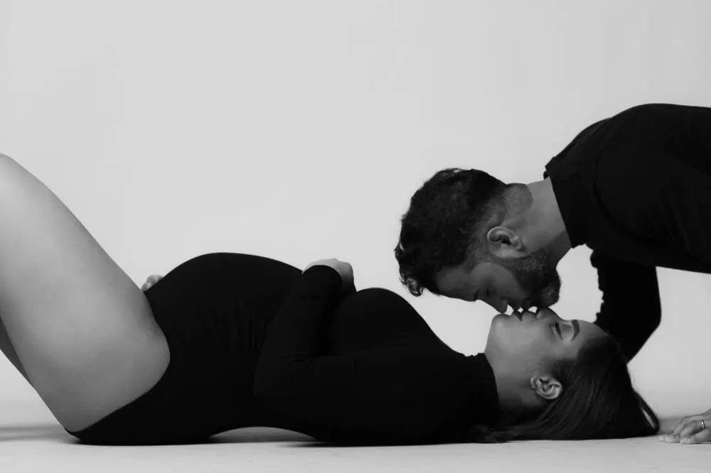Black and white maternity couple portrait on a backdrop, the expecting mother lying back in a black bodysuit as her partner leans down to kiss her, by Create With Amy, a Maryland maternity photographer