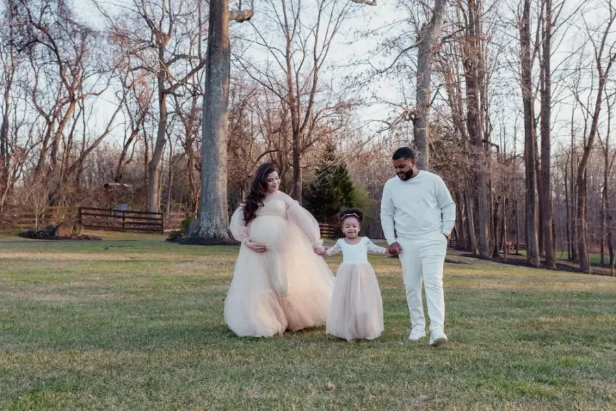 Outdoor maternity photoshoot in an open field, expecting mother in a cream gown walking with her toddler daughter and partner, by Create With Amy