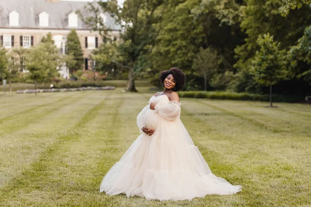 Expecting mother in a cream tulle gown cradling her bump on a wide lawn in front of a manor house, photographed by Create With Amy, Baltimore maternity photographer