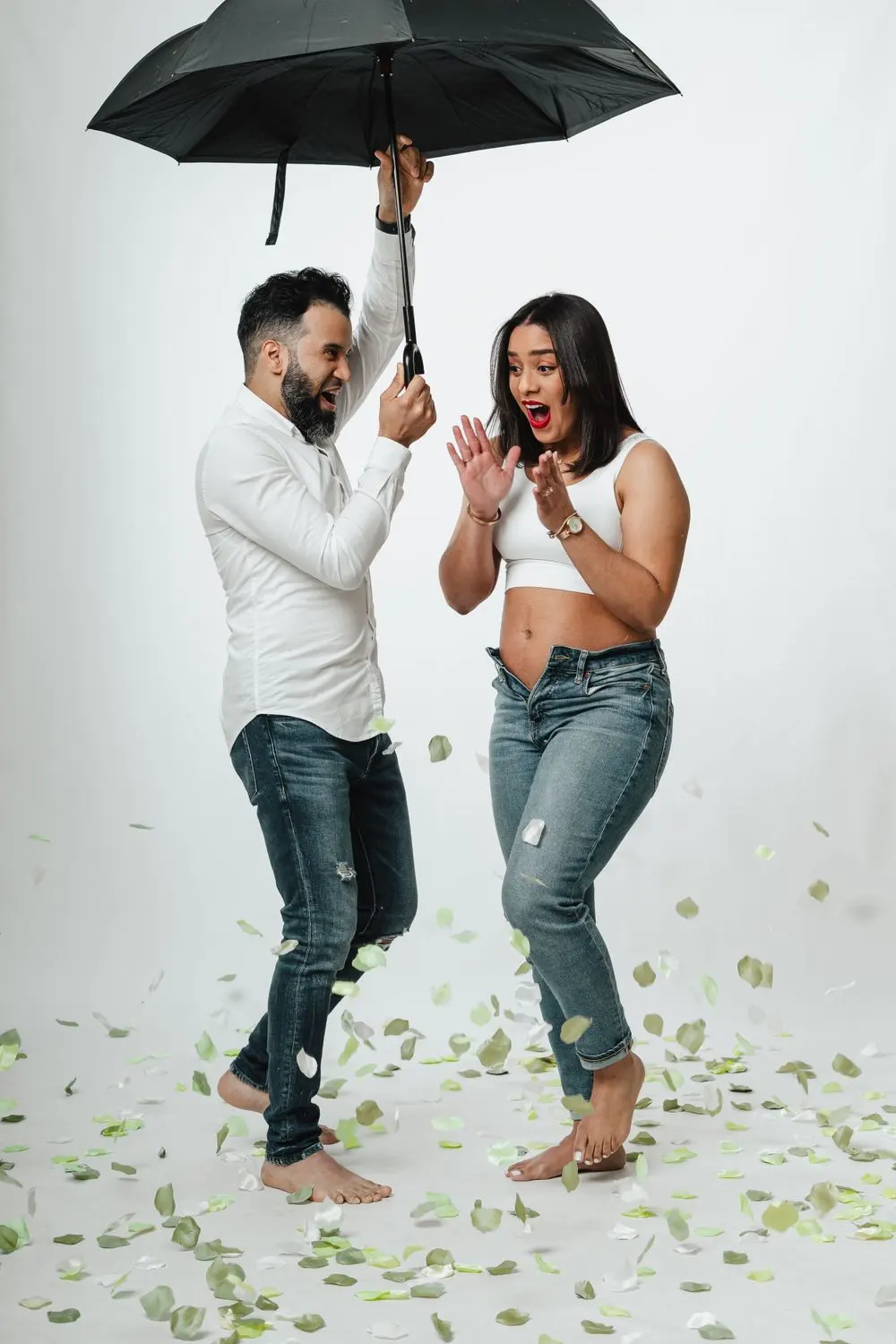 Expecting couple cheering under a black umbrella as green confetti falls on a white backdrop, a gender reveal photographed by Create With Amy